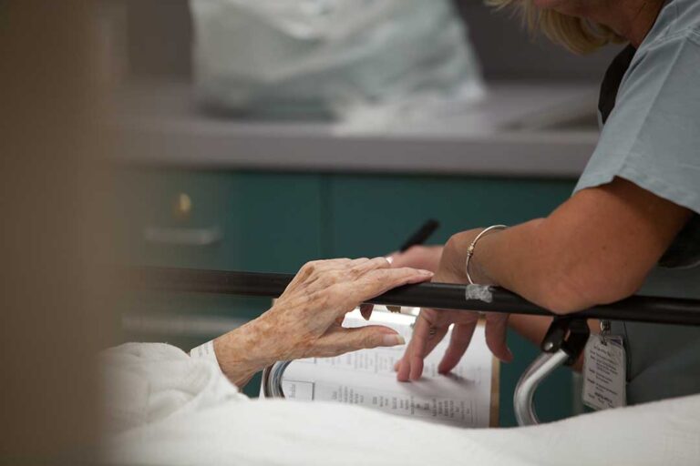 Two older adults and a staff member sitting together as medications and charts are reviewed at a dining table.